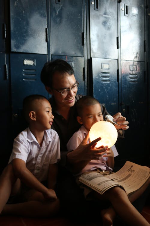 Young smiling ethnic man with decorative lamp and toy plane playing with interested sons in locker room