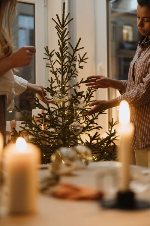 Free Two women decorating a Christmas tree inside a cozy room with candlelight ambiance. Stock Photo
