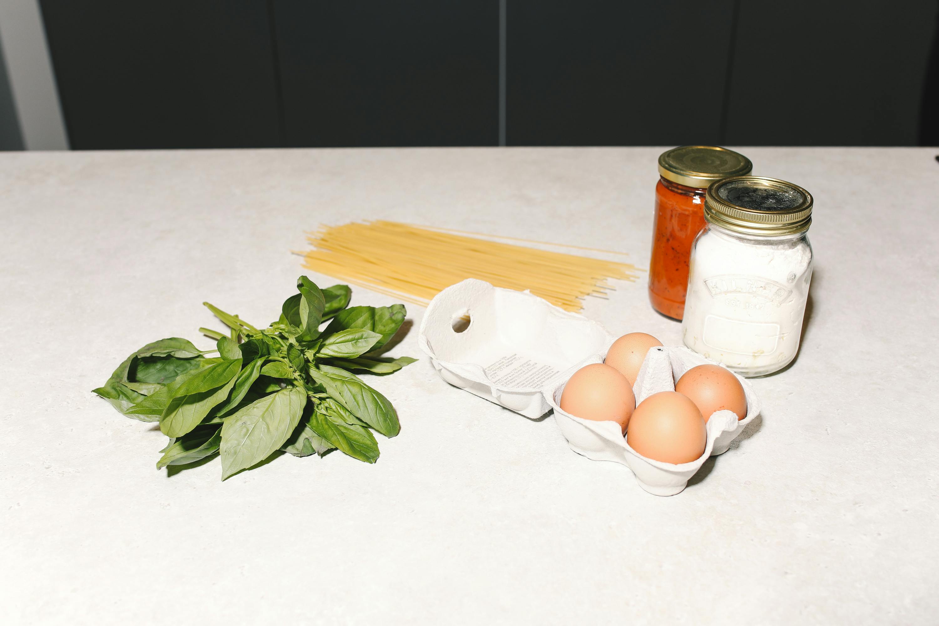Ingredients for homemade pasta with eggs, basil, and jars on a kitchen counter.