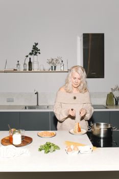Blonde woman cooking spaghetti in a stylish kitchen, enjoying family time.