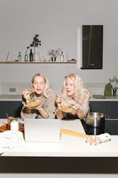 Blonde mother and daughter enjoying pasta at home while watching a laptop in a modern kitchen.