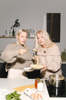 Mother and daughter joyfully cooking spaghetti together in a modern kitchen setting.