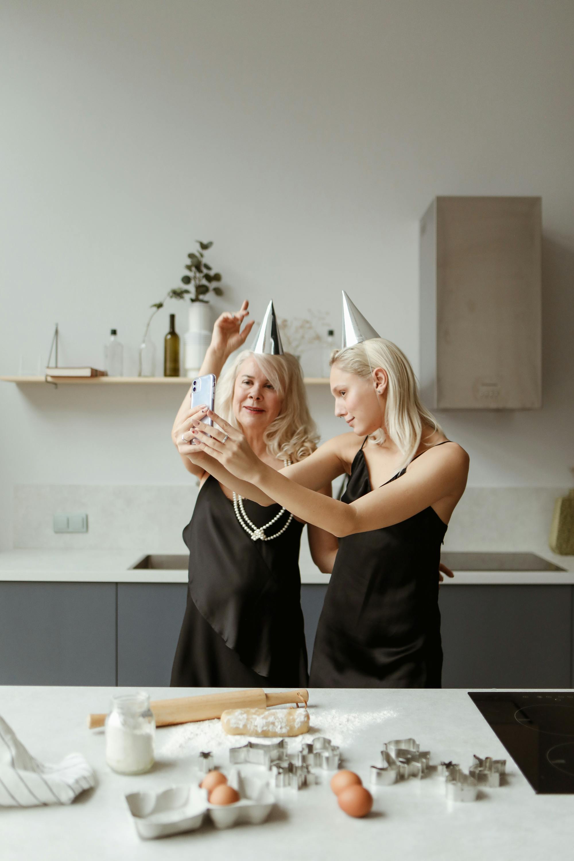 Mother and daughter in black dresses taking a selfie in a modern kitchen, celebrating together.