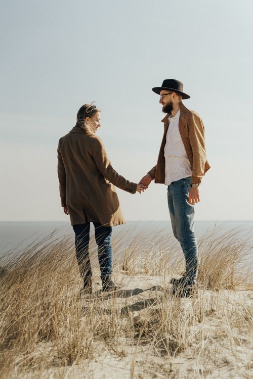Free A couple enjoying a romantic moment by the sea, holding hands on a sandy beach with tall grass. Stock Photo