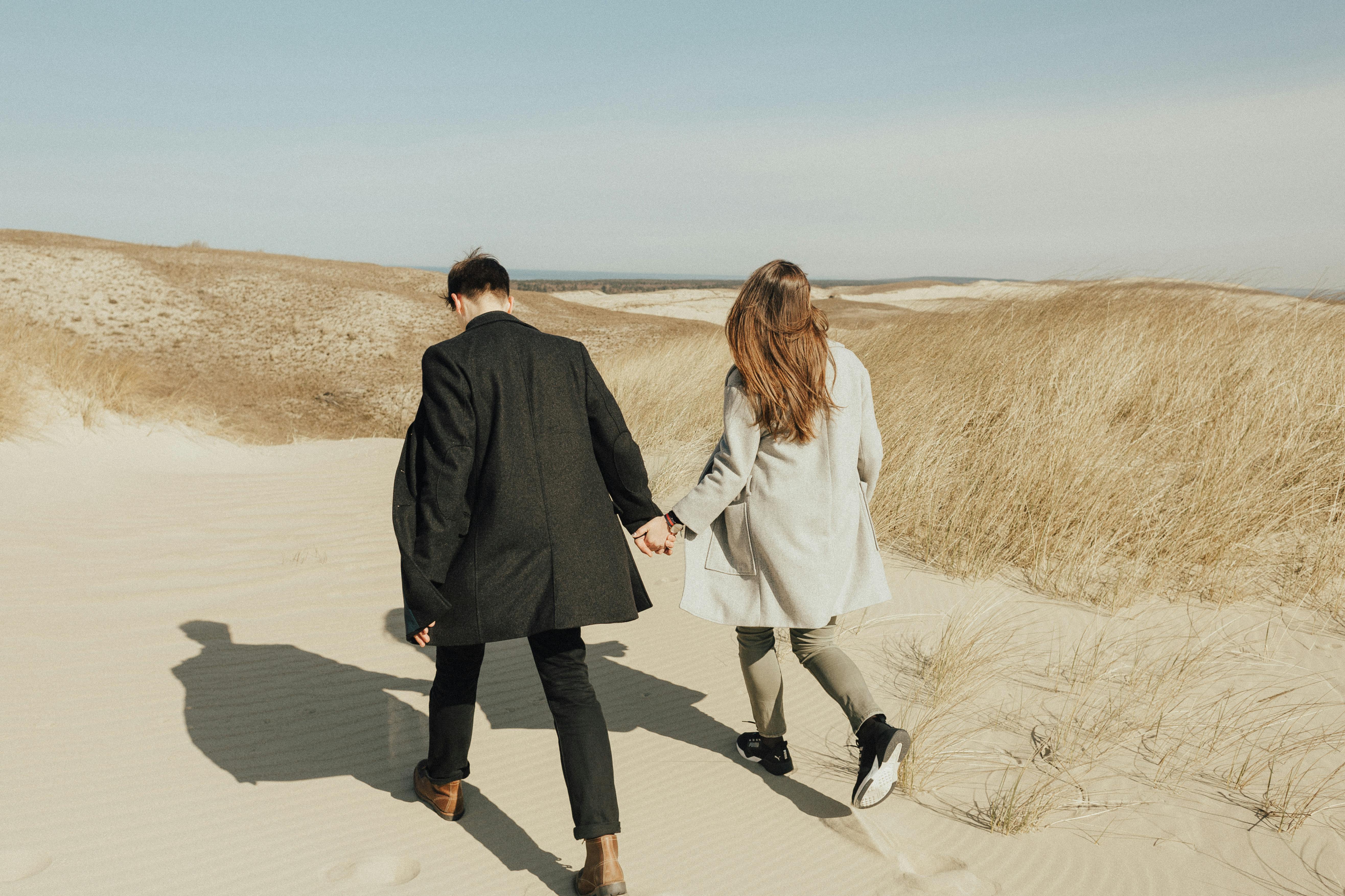 A couple holding hands while walking through sandy dunes under a clear sky.