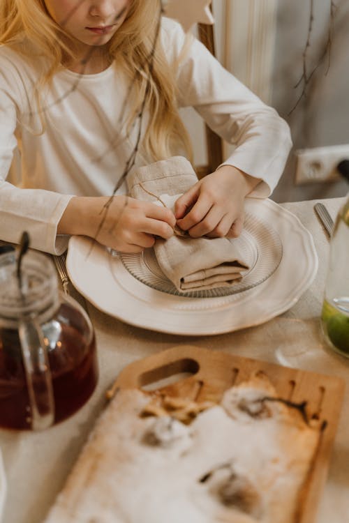 Little Blonde Girl Folding a Napkin at a Dinner Table