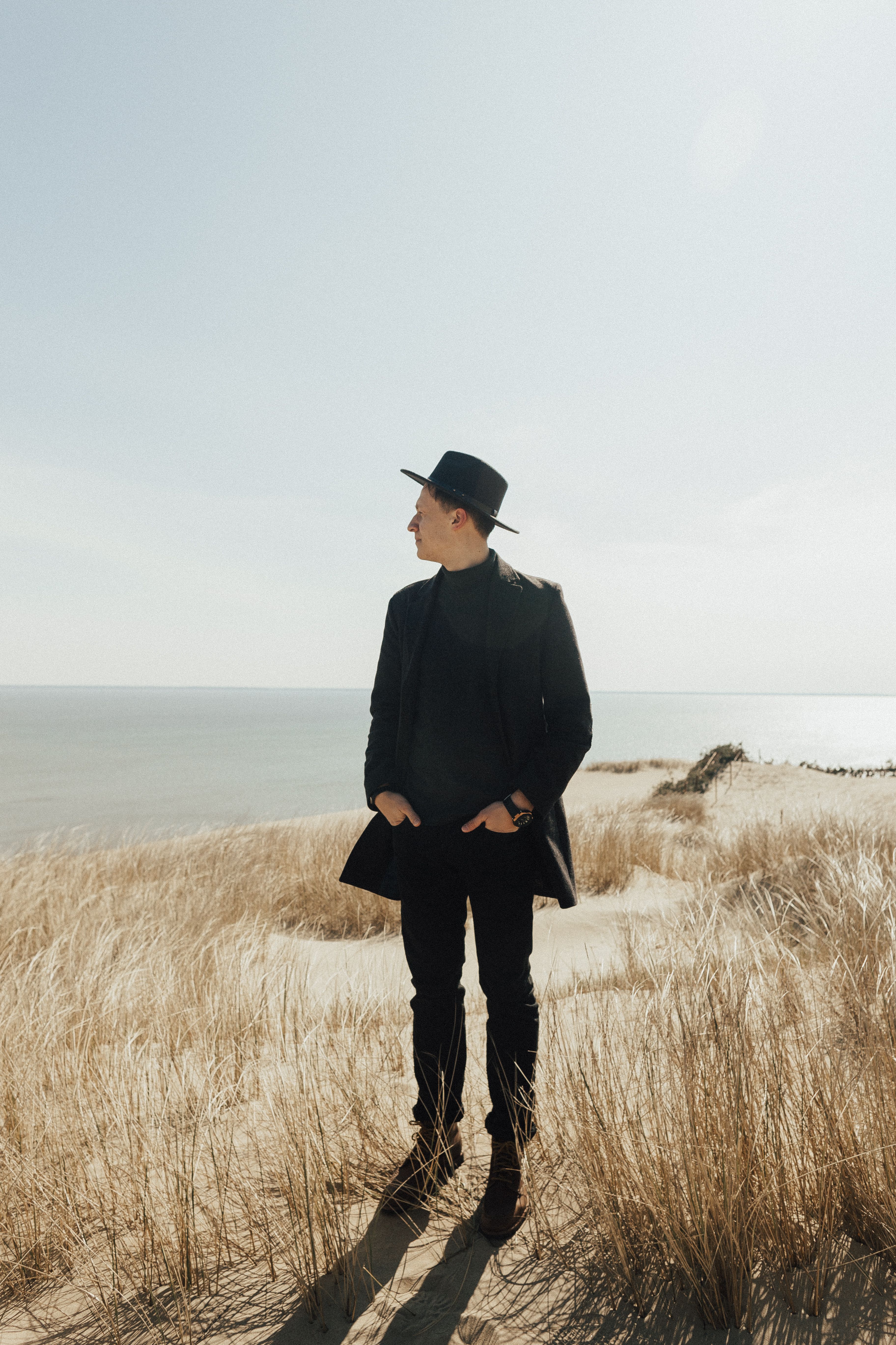 Man in black attire standing on beach dunes with dry grass, exuding elegance and style.
