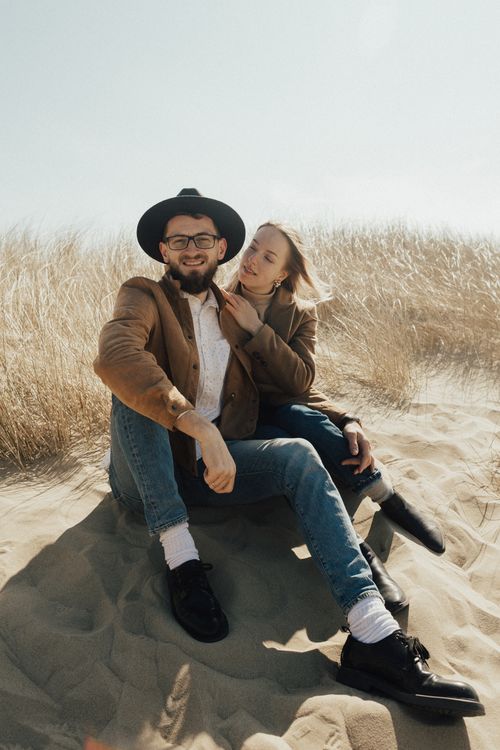 Free A couple sitting on sandy dunes enjoys a bright day in fashionable attire with a natural backdrop. Stock Photo