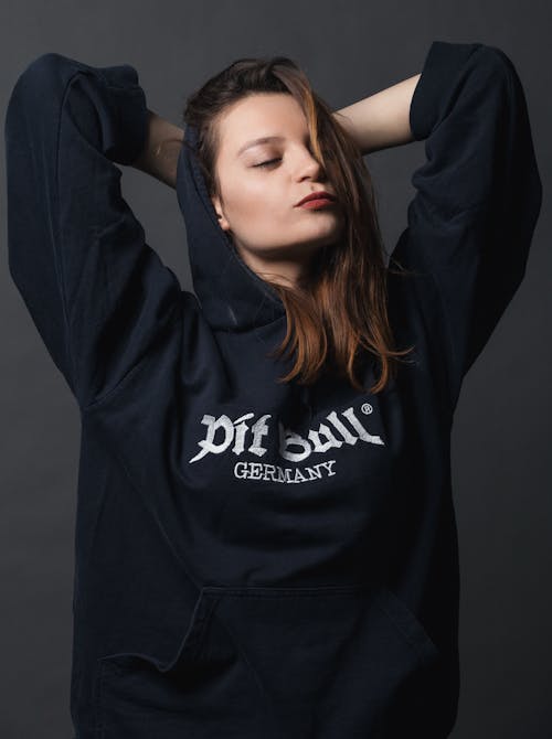 Confident arrogant female wearing black hoodie standing with hands behind head and eyes closed in studio