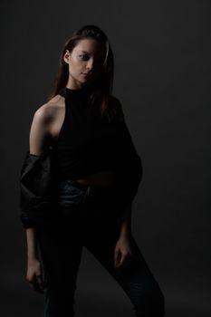 Beautiful emotionless in stylish black clothes and lowered jacket standing against gray wall and looking at camera during photo session in studio