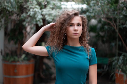 Attractive young female in casual blue top touching curly hair and looking at camera calmly while spending time in lush garden