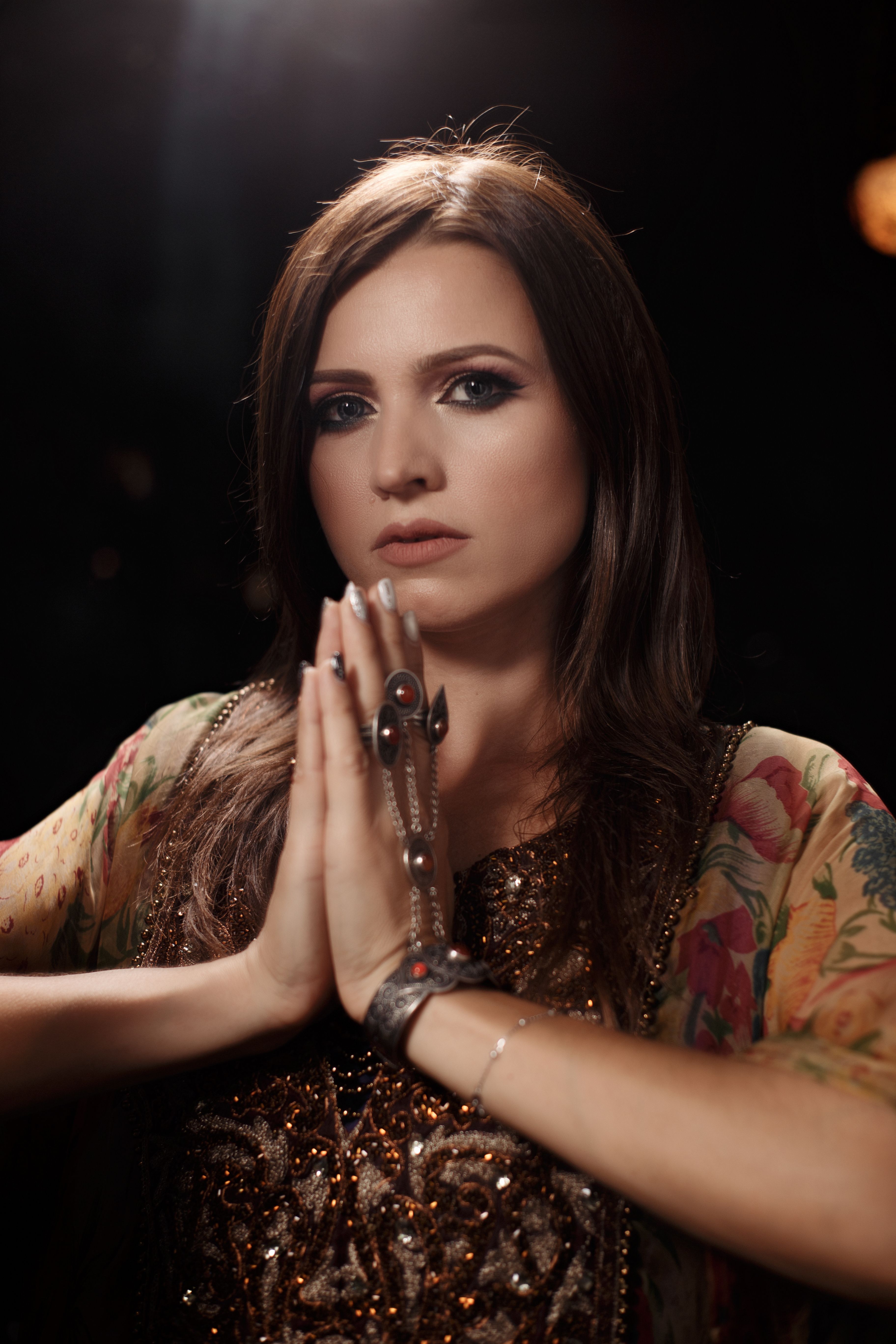 Attractive female wearing rings and bracelets and ornamental dress looking at camera while standing with namaste gesture on black background