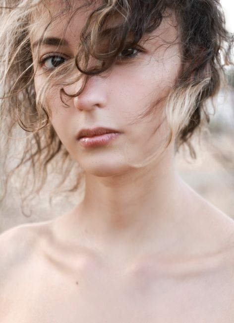Attractive young serious female with bare shoulders looking at camera with thoughtful gaze while standing on blurred background with flying hair in windy weather