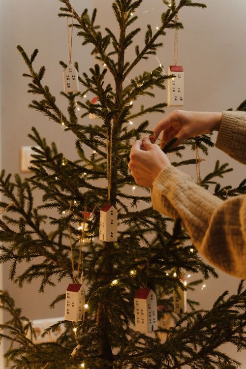 Free Hands adorning a Christmas tree with miniature house ornaments and string lights indoors. Stock Photo