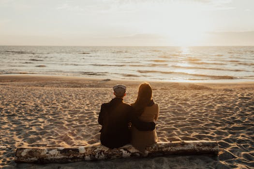 A couple sits together on the beach, embracing as they watch the sunset over the ocean.