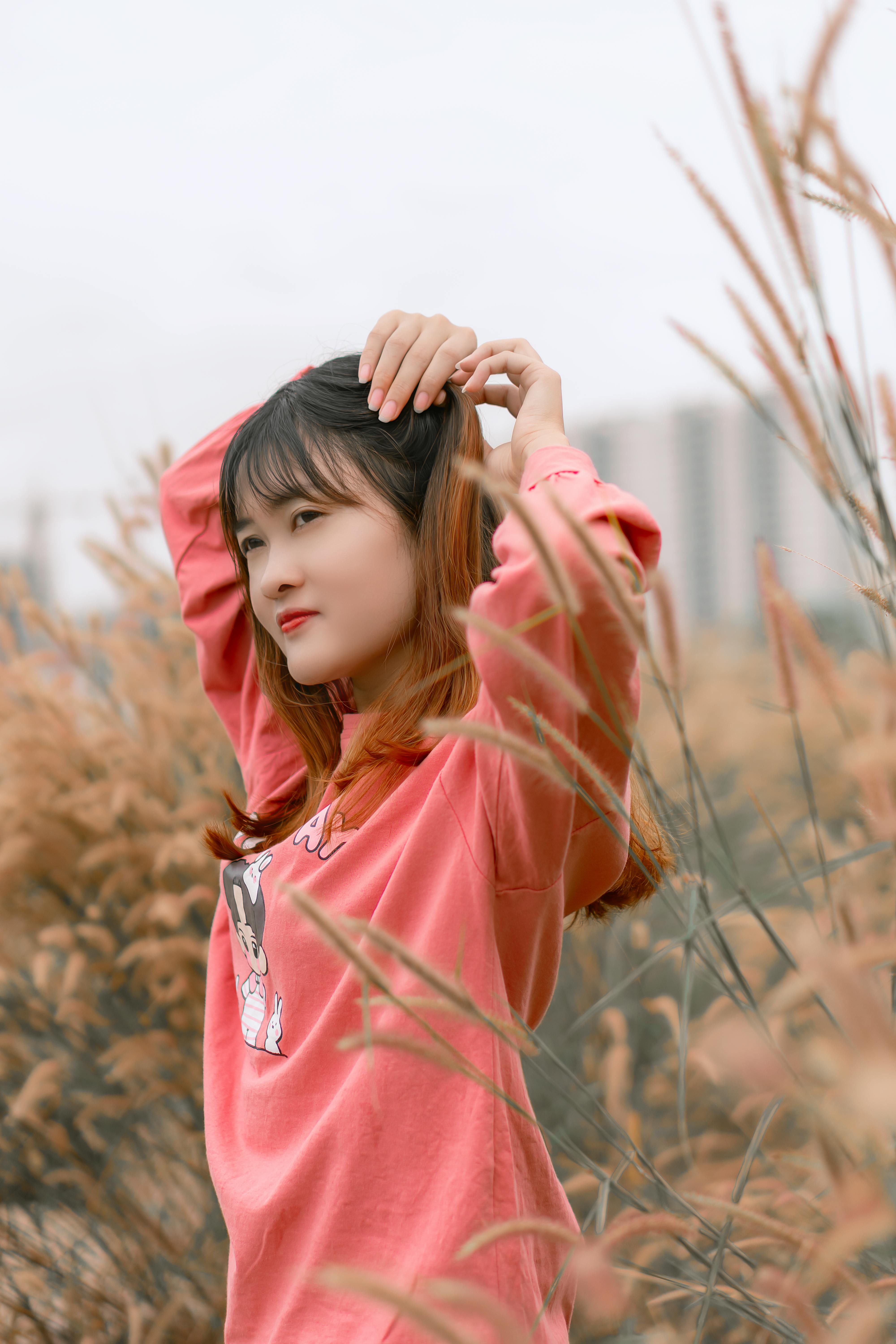 A young woman adjusts her hair outdoors surrounded by tall grass.