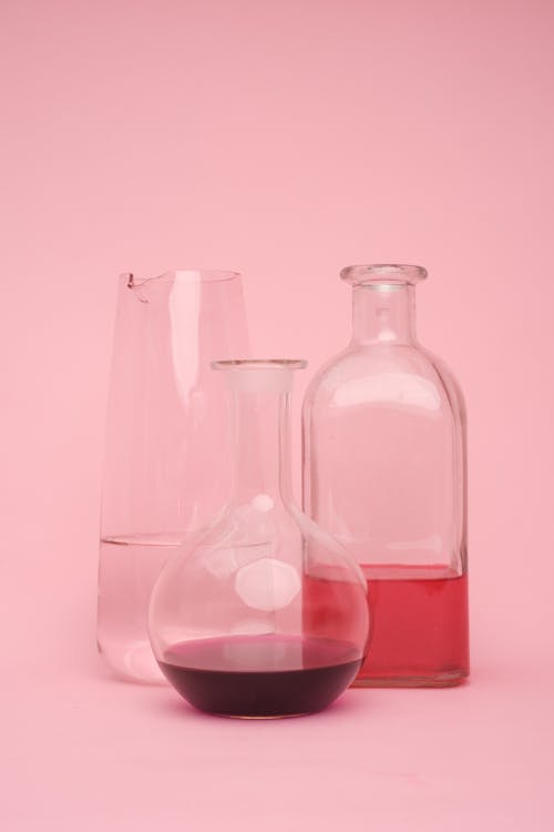 Transparent glass laboratory flasks of various shapes with different chemical liquids placed against pink background