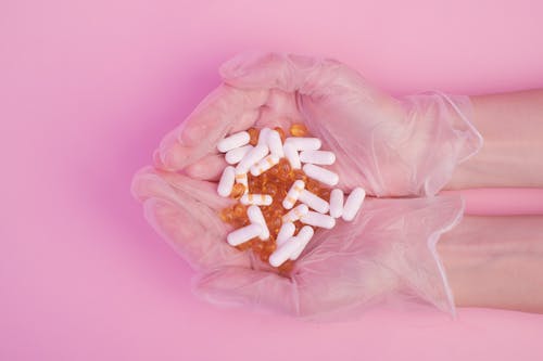From above crop anonymous female in sterile medical gloves with handful of various medicine capsules against pink background