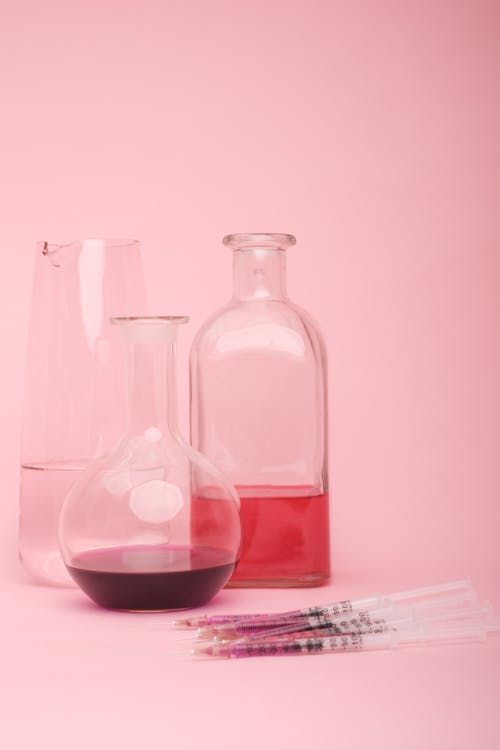 Laboratory glassware of different shapes and set of syringes with various liquids placed against pink background