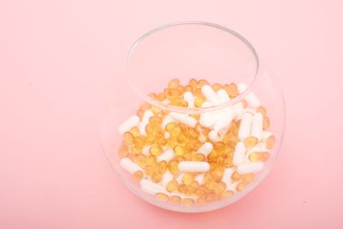 Assorted vitamin capsules in glass bowl on pink surface