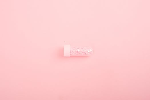 A small test tube filled with glitter lies on a soft pink background, concept of beauty and chemistry.