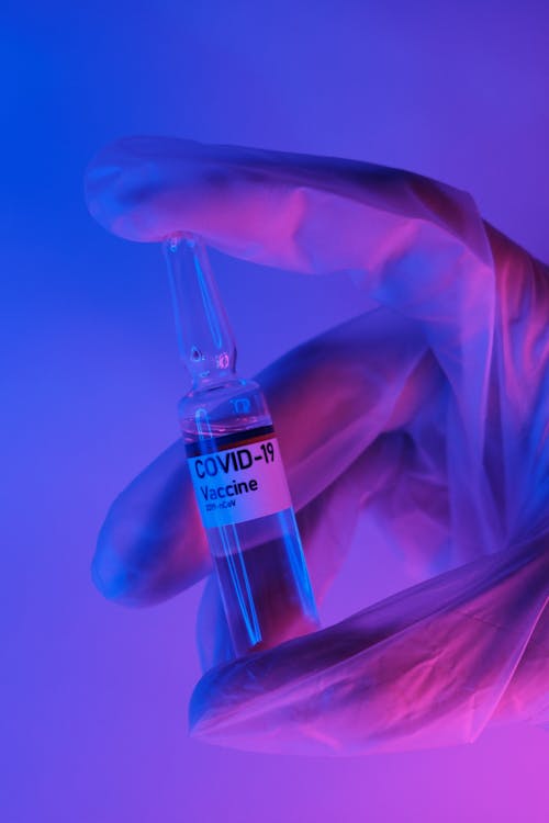 Free Crop unrecognizable medical scientist in protective gloves introducing vial with liquid COVID 19 vaccine in dark laboratory with neon illumination Stock Photo