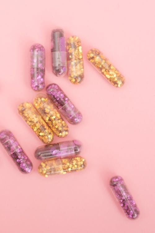 Top view of transparent capsules filled with sparkling purple and golden glitter on pink background