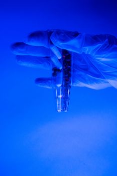 Glass flasks filled with pills and capsules in hand in medical glove in blue neon light
