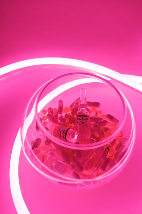 From above of glass bowl filled with pills and ampoules with vaccine for COVID 19 in pink bright neon light