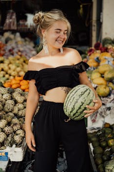 Smiling woman in black attire holds a watermelon at a vibrant market in Bali, Indonesia.