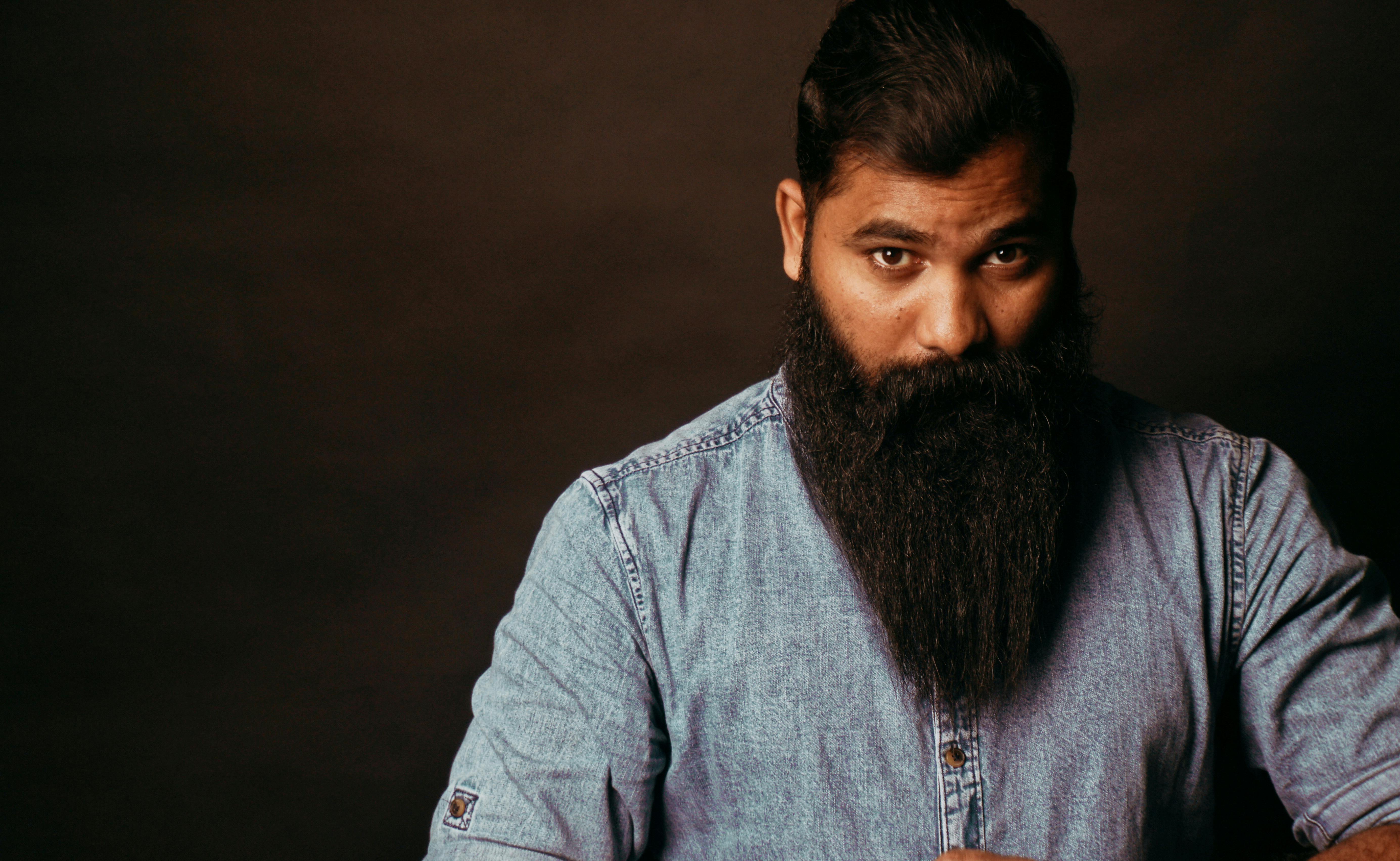 A striking portrait of a man with a long beard wearing a denim shirt, set against a dark background.