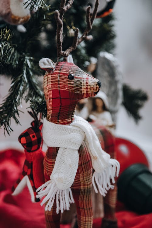 Cute handmade checkered reindeer with scarf hanging on branch of Christmas tree in room on blurred background during holiday celebration