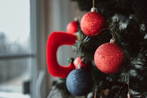 Red and blue decorative baubles hanging on branches of Christmas tree near window on blurred background during holiday preparation in apartment
