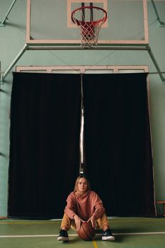 A young woman sits on an indoor basketball court holding a ball, under a hoop.