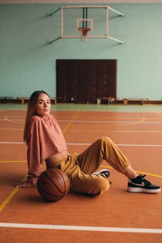 A young woman sits casually on a basketball court, exuding confidence and style.