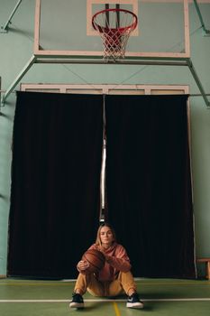 Young woman sitting on an indoor basketball court holding a ball under the hoop.