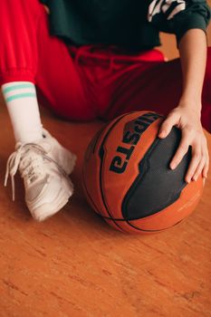 A person in jogger pants sitting on a wooden floor holding a basketball.
