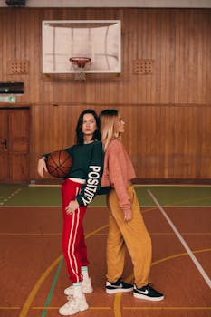 Two women in casual outfits pose together on an indoor basketball court holding a ball.