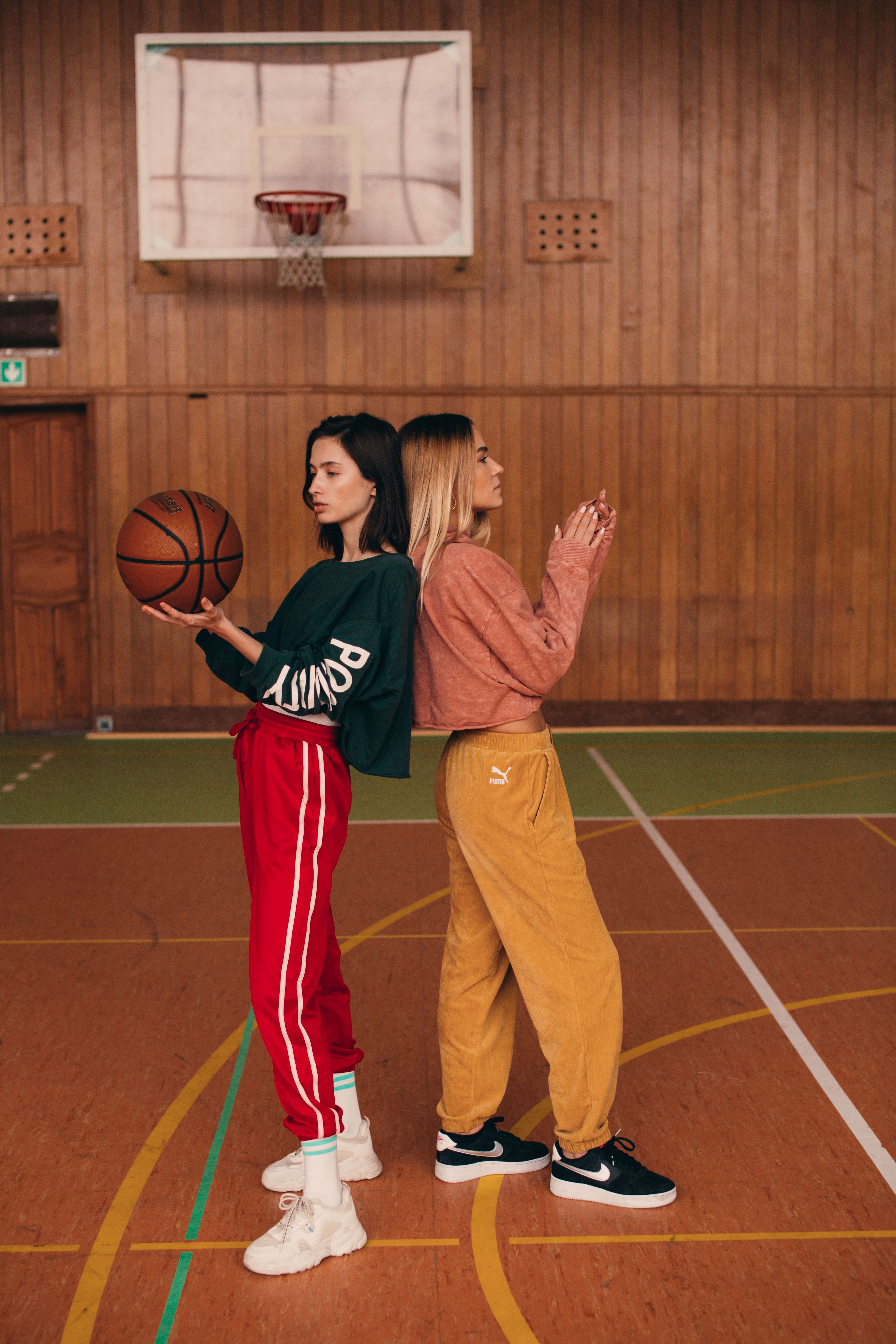 Two young women standing back to back on an indoor basketball court, one holding a ball.