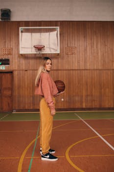 Side view of a woman holding a basketball in a wooden-paneled indoor court wearing casual attire.