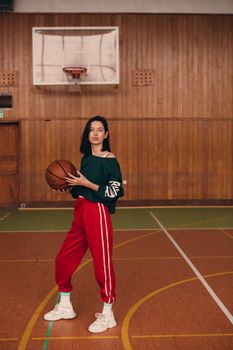 A fashionable young woman posing confidently with a basketball inside a gymnasium.