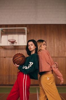 Two women posing with a basketball in a gym, showcasing casual athletic fashion.