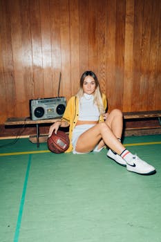 A stylish young woman sits with a basketball and boombox, showcasing retro fashion in a gym setting.
