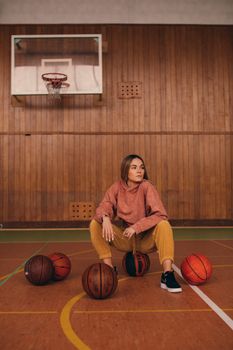 Woman sitting surrounded by basketballs on an indoor court, wearing a hoodie.