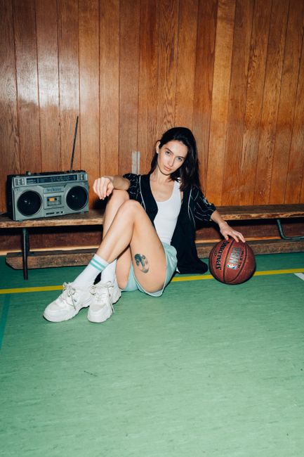 Full body of confident female looking at camera while sitting on green sports ground with crossed bent legs near wooden wall and record player
