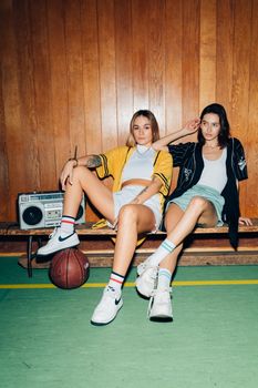 Two stylish women seated in a gym, exuding modern urban fashion with a vintage vibe.