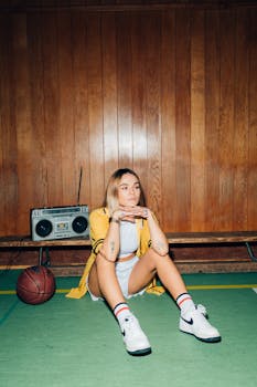 Portrait of a young woman in a gym, sitting with a basketball and vintage boombox.