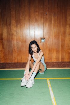 Woman in casual attire sitting on a gym floor against a wooden wall.