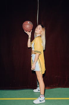 Stylish young woman in a yellow jacket poses confidently with a basketball against a dark background.