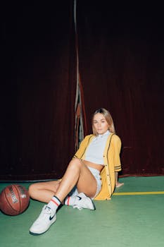 Stylish young woman in yellow jacket posing with basketball in an indoor court.